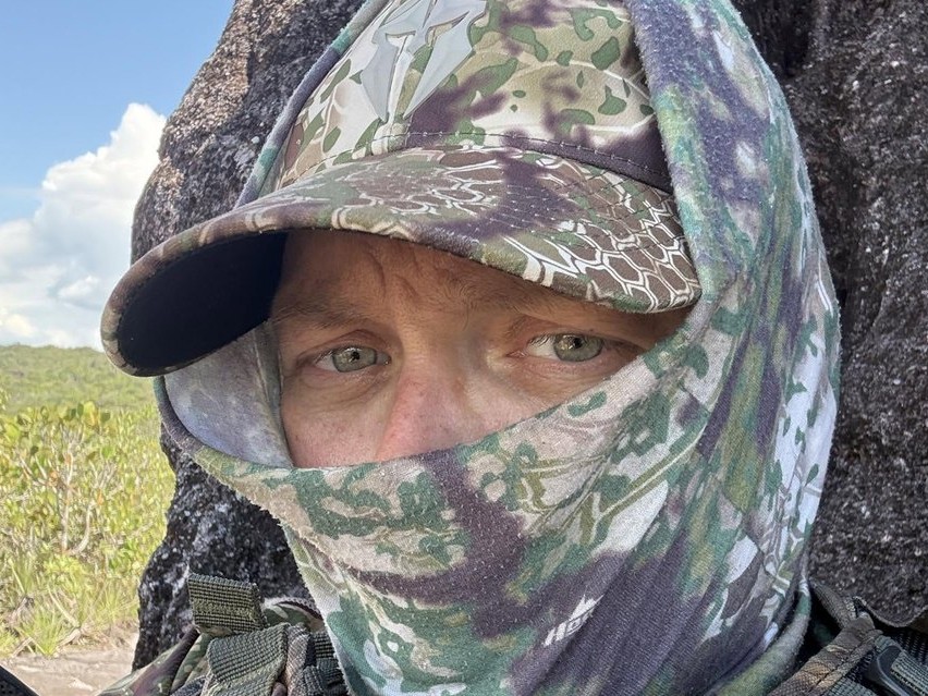 Adam Rainoff con ropa de camuflaje junto a una roca